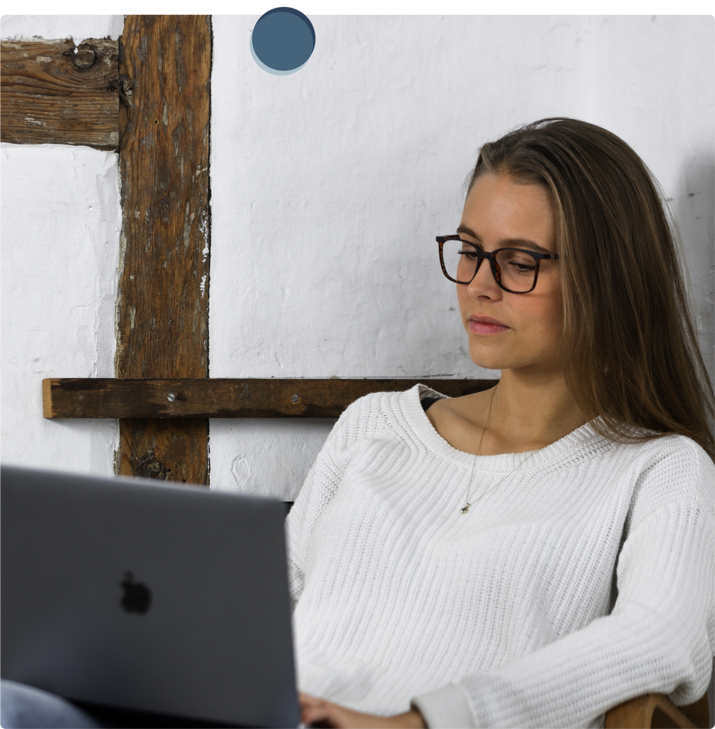 Eine junge Frau mit weißem Pullover trägt eine Blaufilter Brille, während Sie auf Ihren Laptop schaut